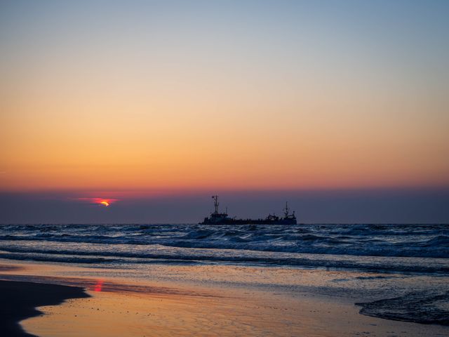 Orange and pink sunset reflected on wet Baltic sand at Łeba, with a ship silhouetted near the horizon.