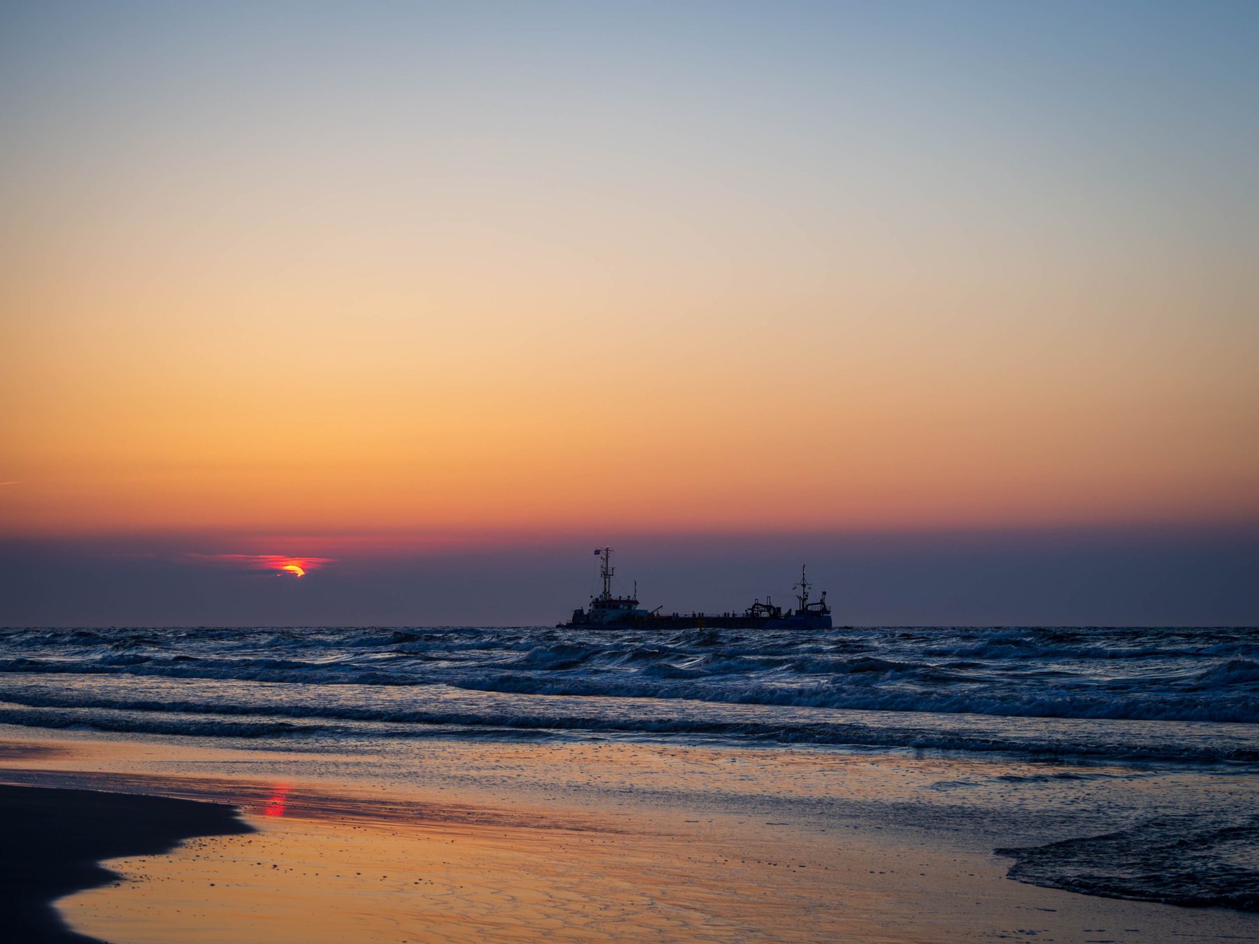 Orange and pink sunset reflected on wet Baltic sand at Łeba, with a ship silhouetted near the horizon.