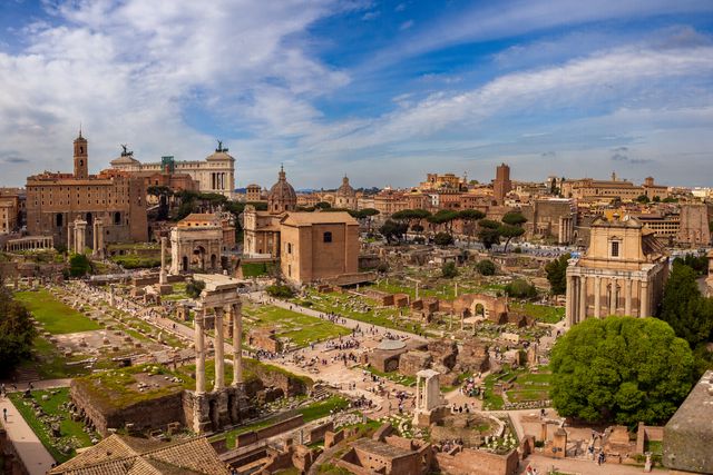 Panoramic view across the Roman Forum, with columns, ruins, trees, and ochre buildings under blue sky.