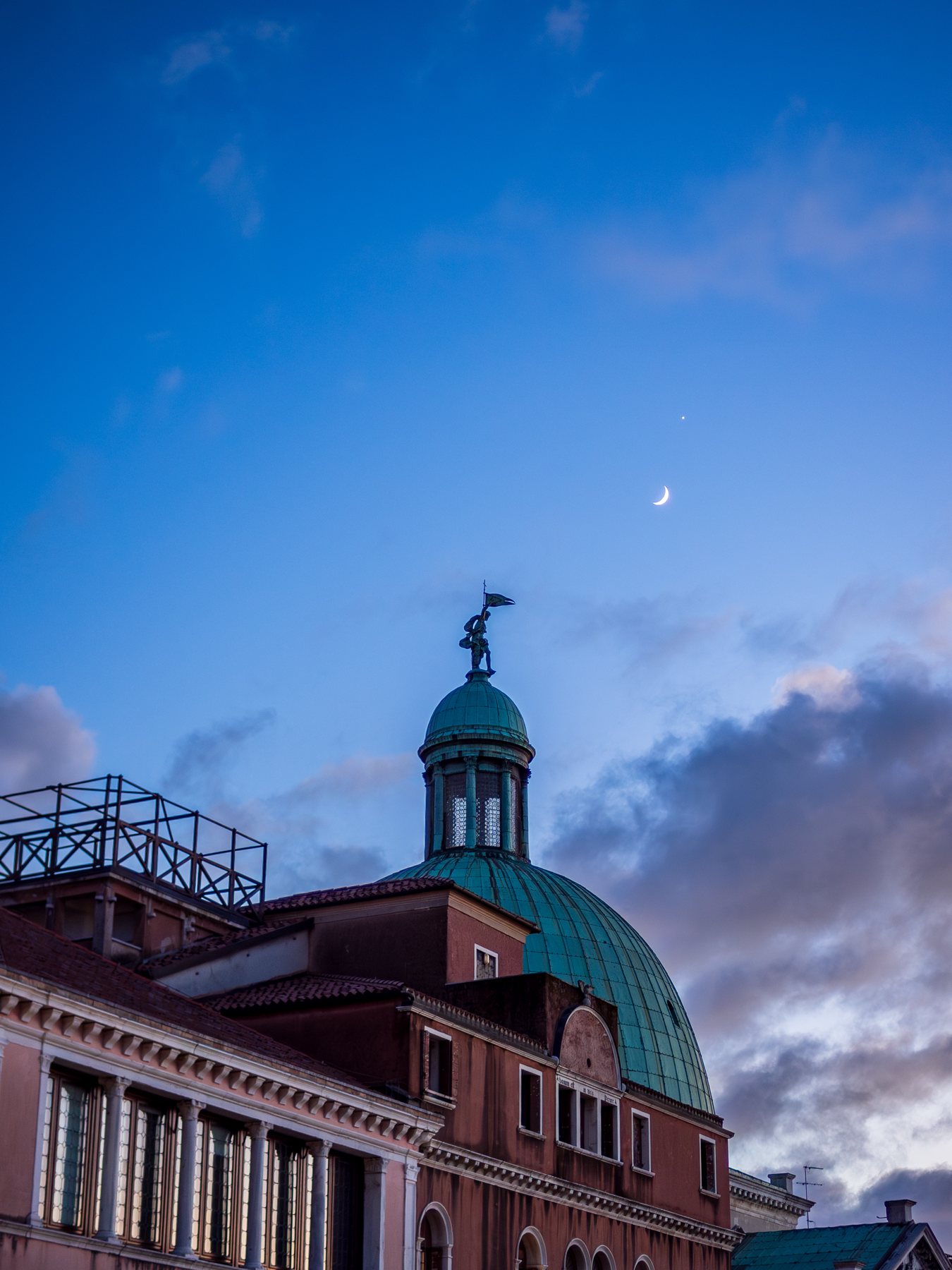 A slim crescent moon above a green copper dome and pink clouds at winter dusk in Venice.