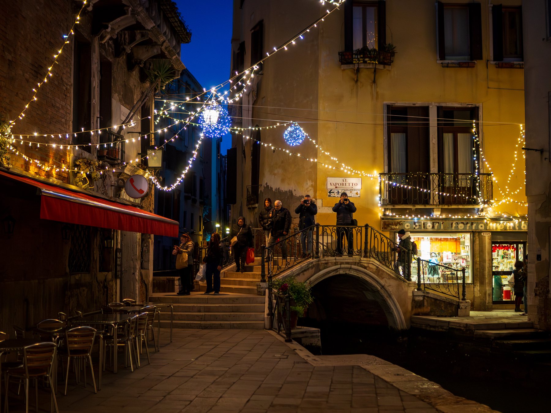 Festive lights leading down a warm Venetian street toward a small arched bridge at night.