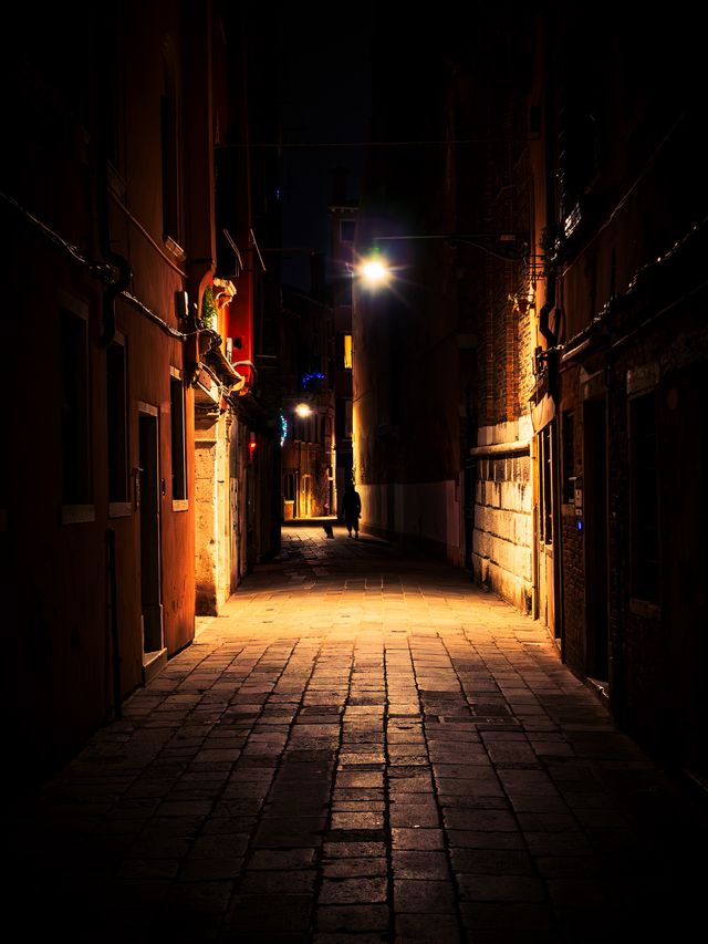 A narrow stone lane in Venice disappearing between dark walls toward a single warm streetlight.