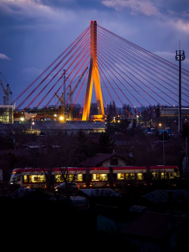 Gdańsk's cable-stayed bridge illuminated orange and violet above a train at blue hour.