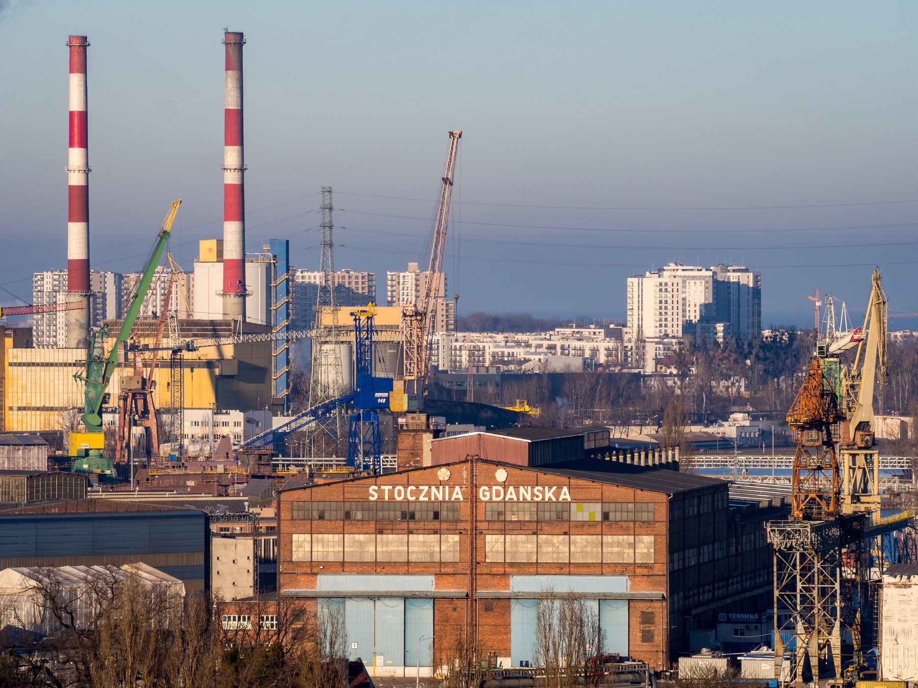 Shipyard cranes, striped chimneys, and the historic Stocznia Gdańska workshop in clear winter light.
