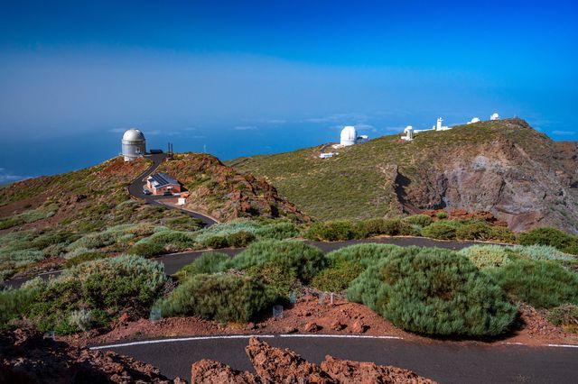 Observatory domes among rounded green shrubs and red volcanic paths on La Palma's summit ridge.
