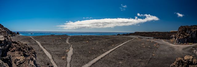 Panoramic road through black volcanic terrain descending toward the Atlantic and a distant island.