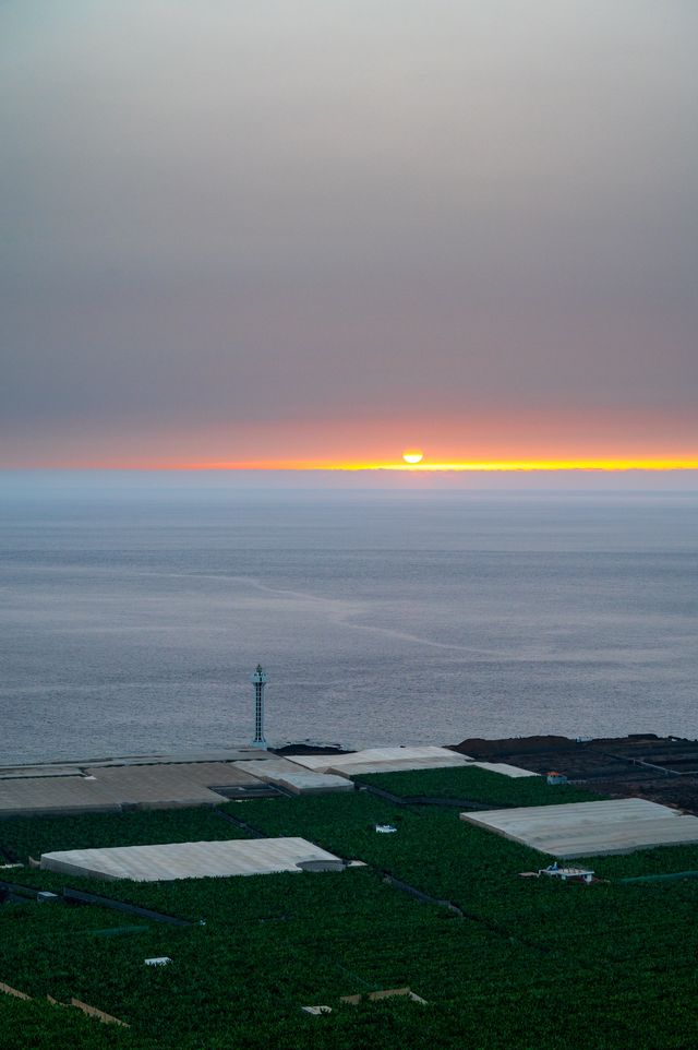 The sun touching the Atlantic horizon beyond banana fields and a slender coastal lighthouse.