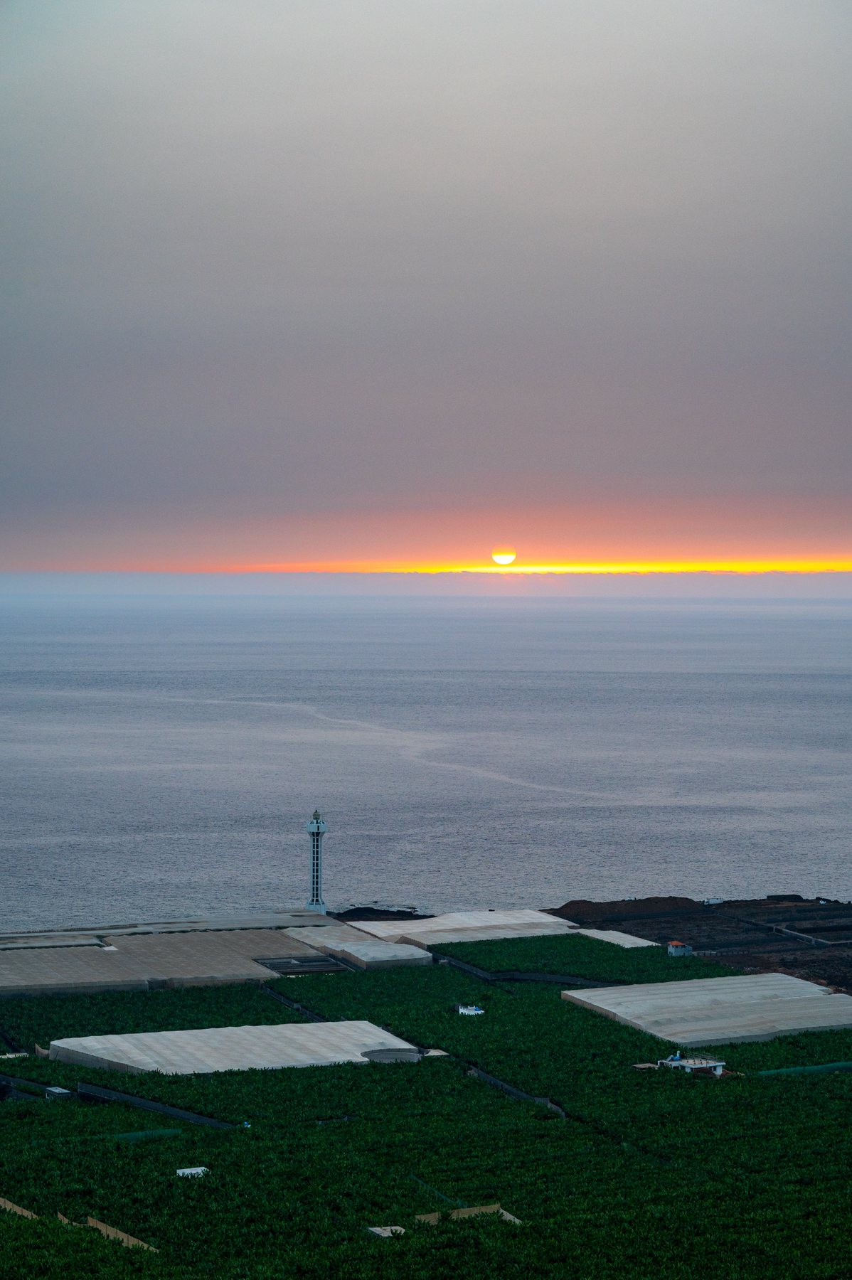 The sun touching the Atlantic horizon beyond banana fields and a slender coastal lighthouse.