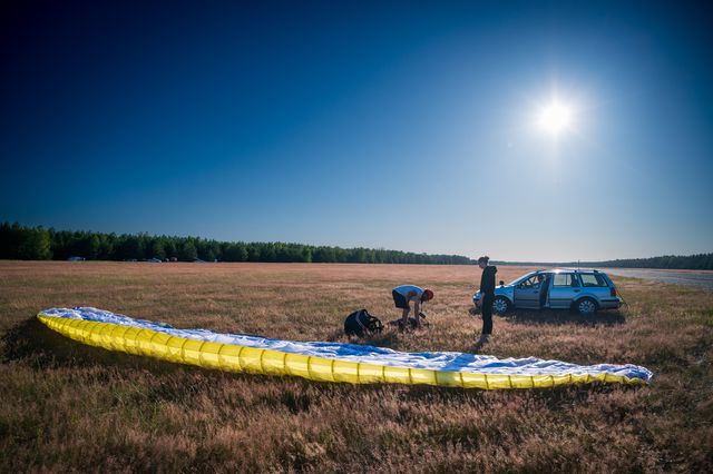 A yellow paraglider canopy laid across dry grass as pilots prepare beside a parked car.