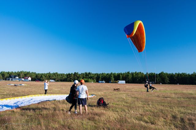 An orange paraglider rising above people and laid-out wings on the open airfield.