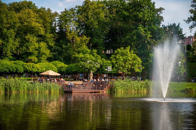 A lake fountain and tree-lined outdoor seating establishing the summer concert setting in Lidzbark.