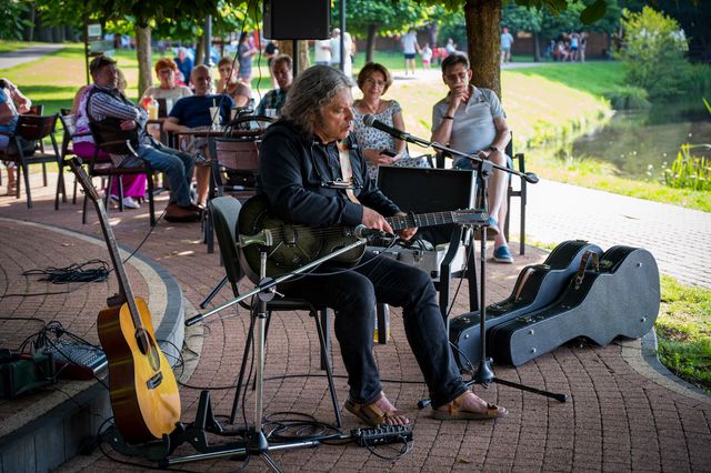 Paweł Szymański seated with guitar and microphone before a small audience beside the lake.