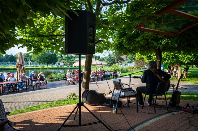 View from behind the outdoor stage toward listeners seated under trees in Lidzbark.