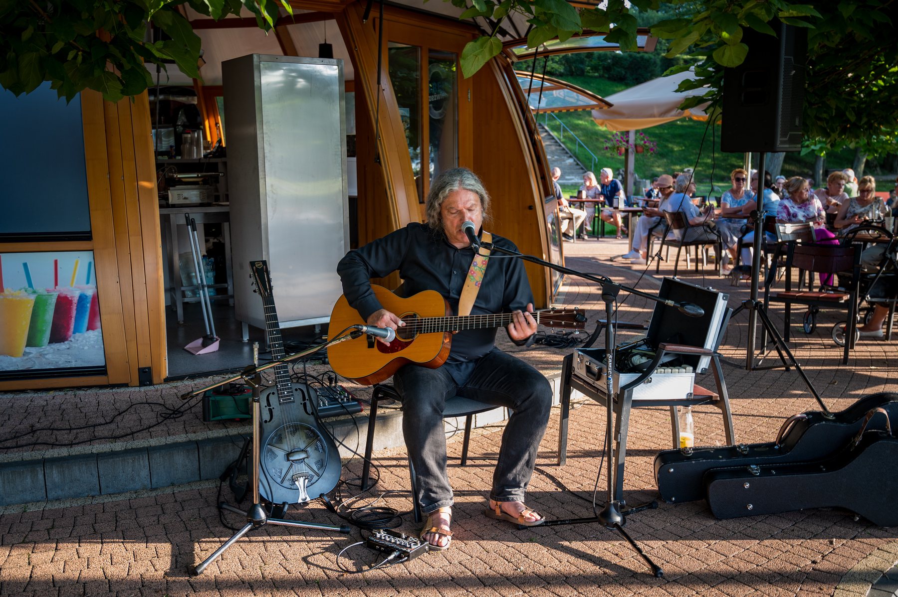 Paweł Szymański singing and playing acoustic guitar on an intimate park stage.