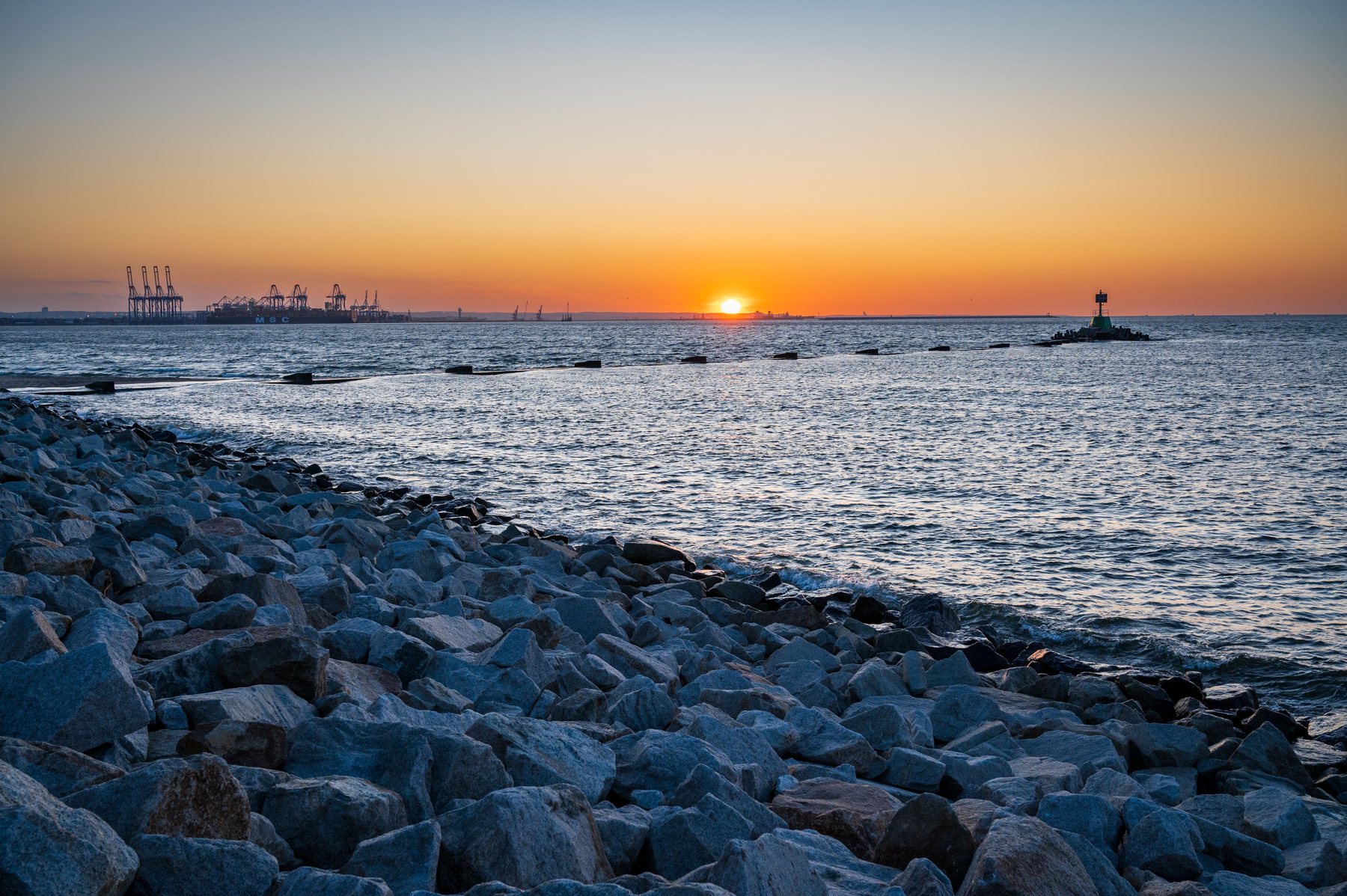 Sunset at the end of a rock breakwater, with port cranes and a navigation beacon on the horizon.