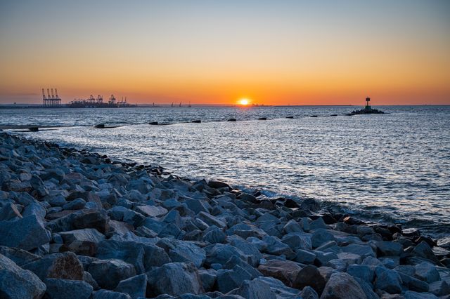 Sunset at the end of a rock breakwater, with port cranes and a navigation beacon on the horizon.