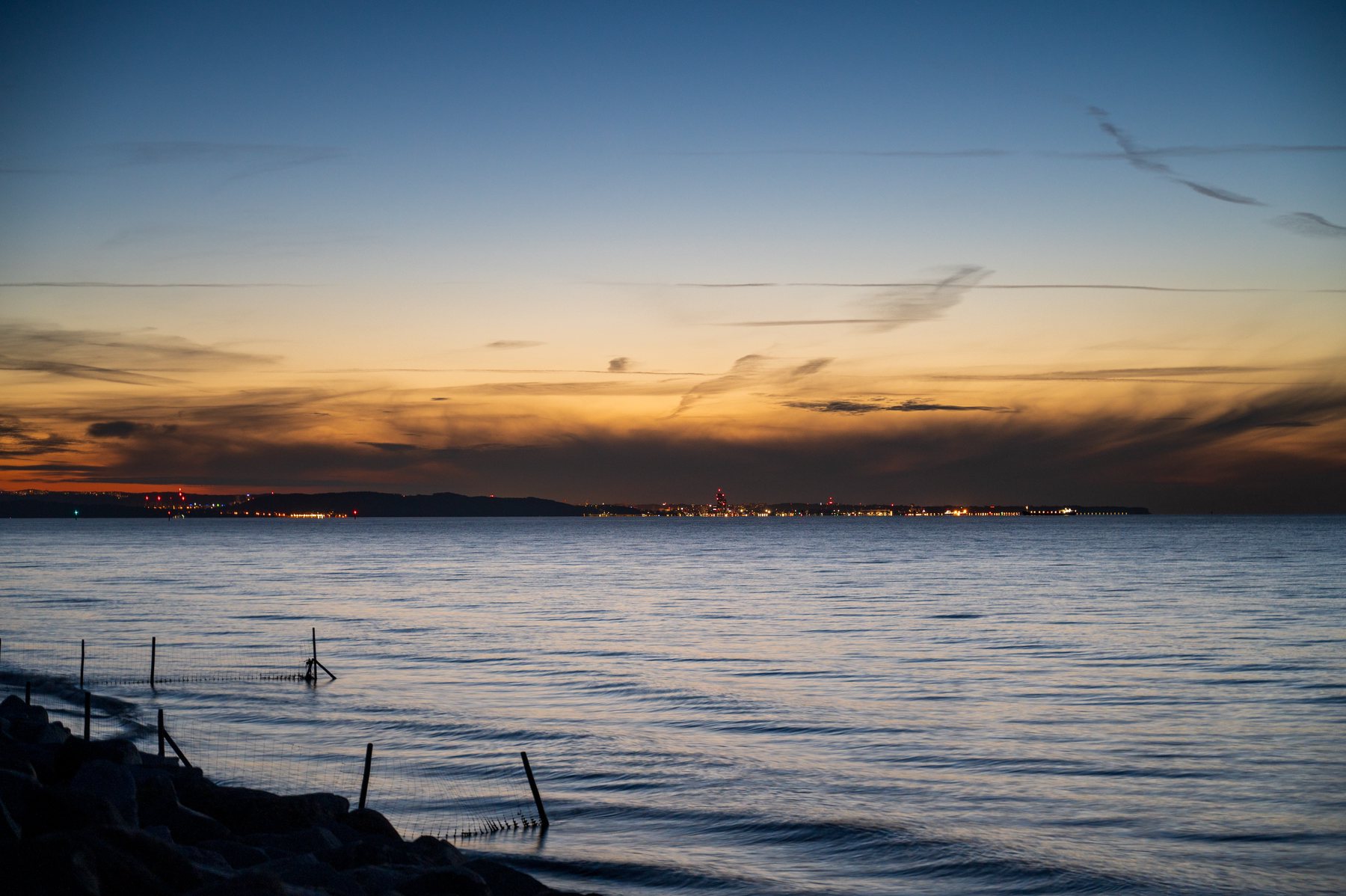 Quiet blue-hour water beneath delicate clouds and a thin band of orange city lights.
