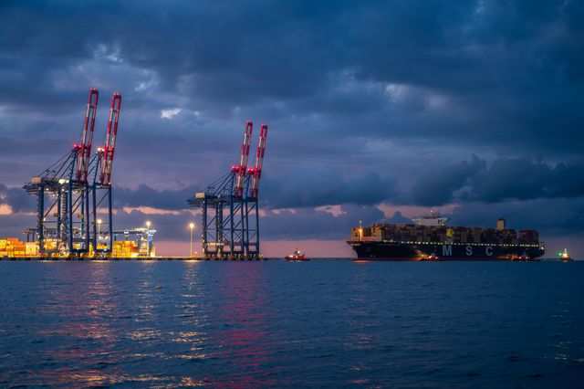 Illuminated container cranes and a large cargo ship beneath dark blue storm clouds.
