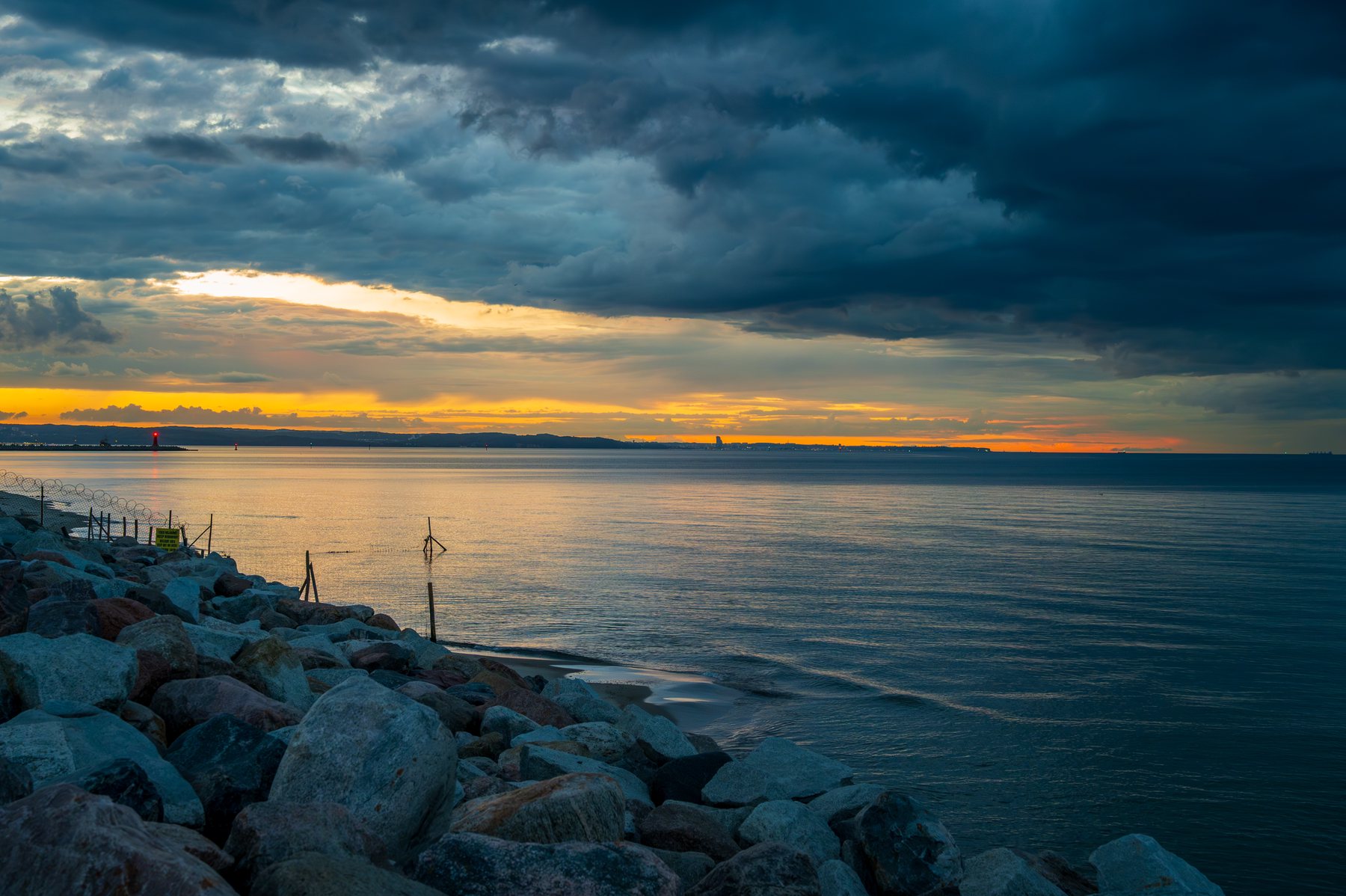 A narrow yellow sunset opening beneath heavy blue clouds above a calm rocky shoreline.