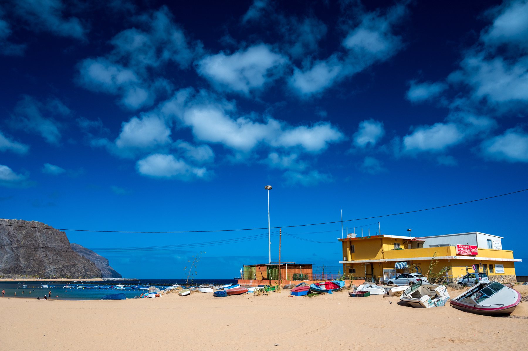 Small fishing boats pulled onto sand beside a yellow waterfront restaurant and blue Tenerife cove.