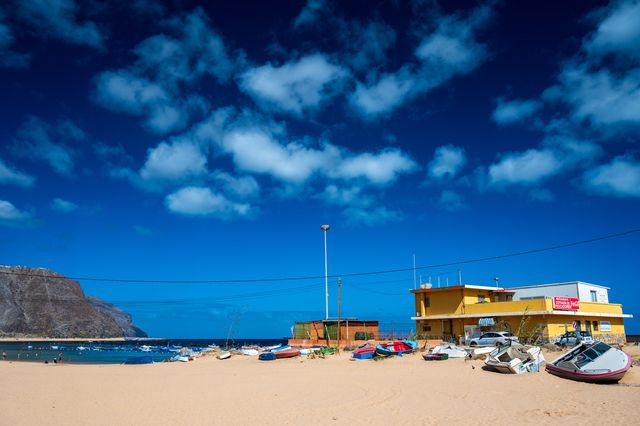Small fishing boats pulled onto sand beside a yellow waterfront restaurant and blue Tenerife cove.