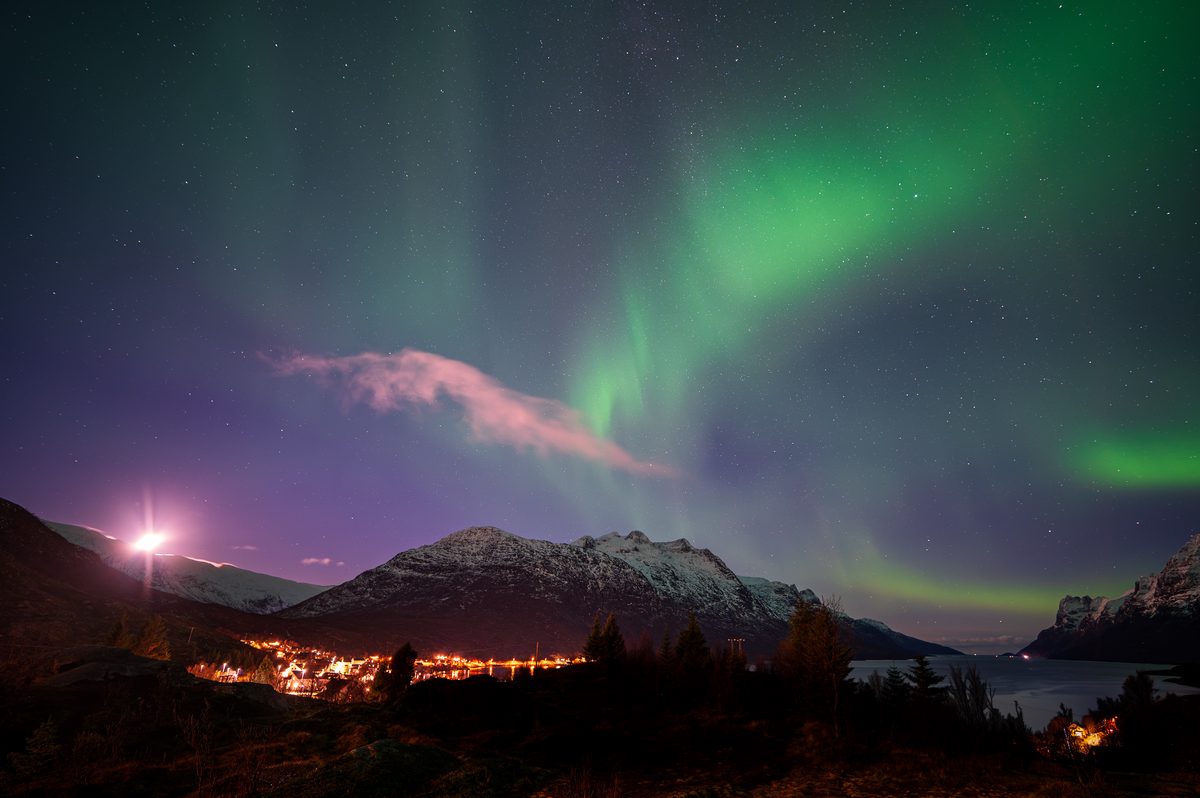 Vibrant green aurora borealis illuminates a snow-covered mountain valley with glowing village lights below, creating a magical Arctic night scene.