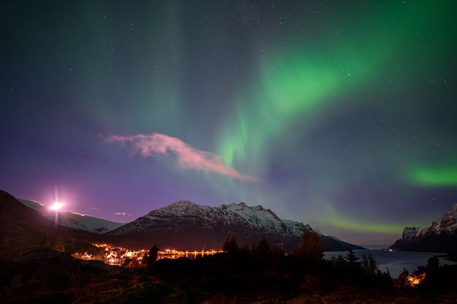 Vibrant green aurora borealis illuminates a snow-covered mountain valley with glowing village lights below, creating a magical Arctic night scene.