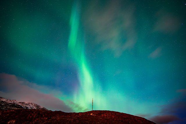 Vibrant northern lights dance across the Arctic sky above a snow-covered mountain ridge with an illuminated radio tower.