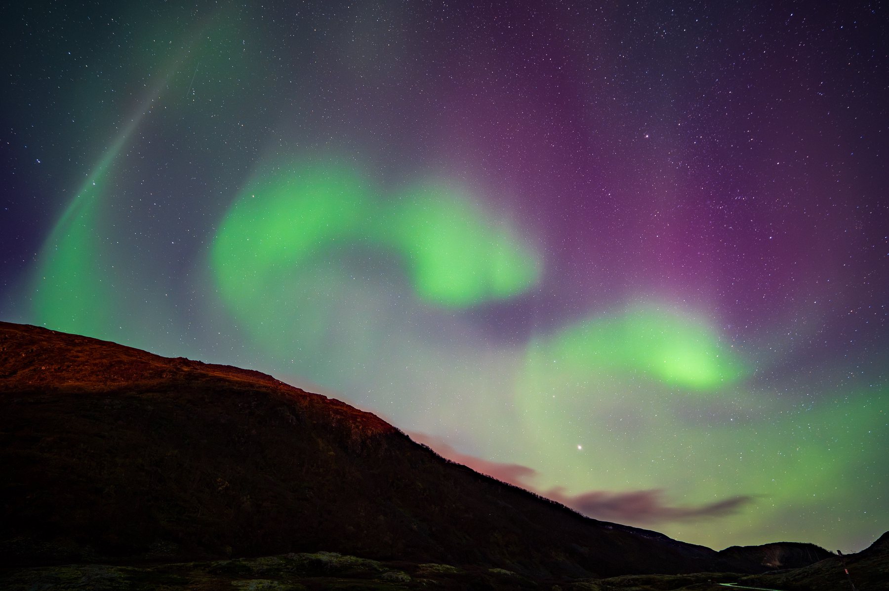 Vibrant green and purple aurora borealis dancing across the night sky above a dark mountainous landscape, with stars twinkling overhead.