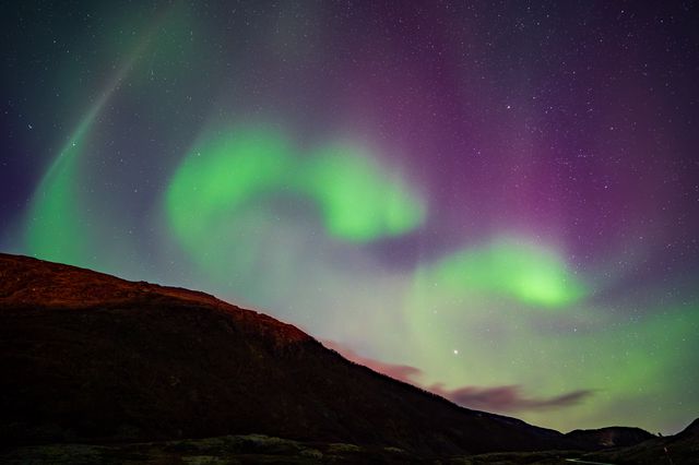 Vibrant green and purple aurora borealis dancing across the night sky above a dark mountainous landscape, with stars twinkling overhead.
