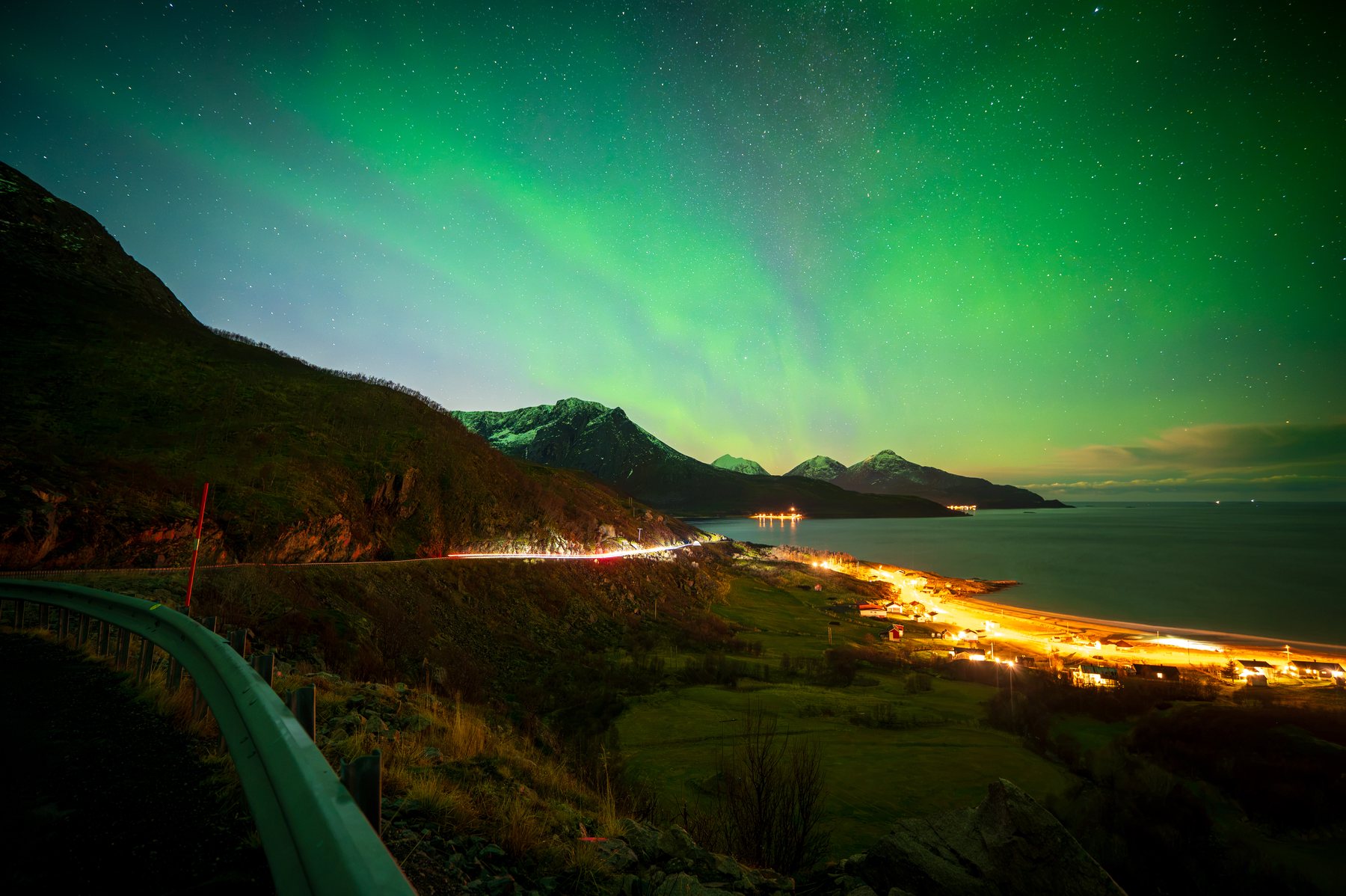 Ethereal northern lights dance above a coastal mountain village, casting an otherworldly green glow over illuminated seaside settlements and dark valleys.