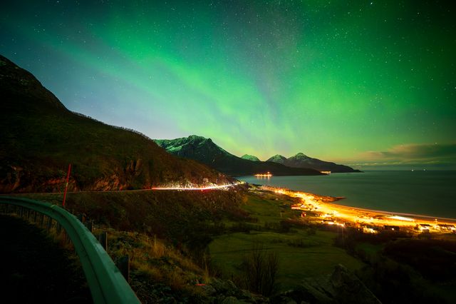 Ethereal northern lights dance above a coastal mountain village, casting an otherworldly green glow over illuminated seaside settlements and dark valleys.