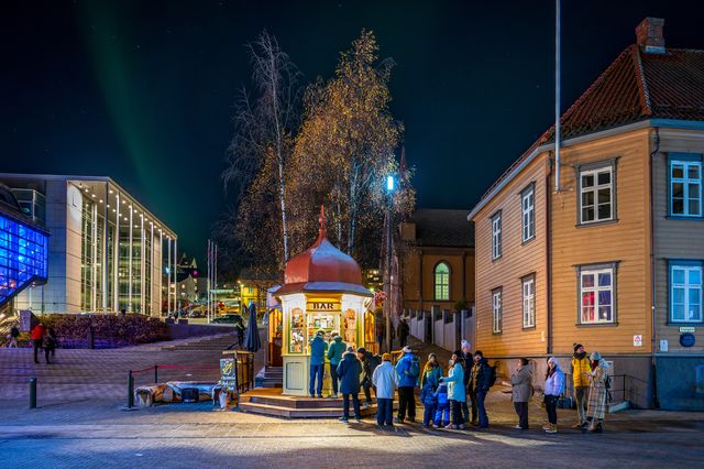People gathered at a warmly lit kiosk on a Tromsø street while faint aurora glows overhead.