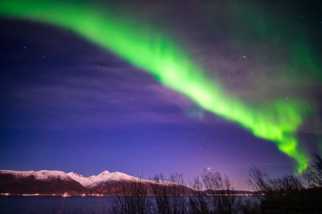 A sweeping green aurora arc above snowy mountains reflected faintly in dark water.