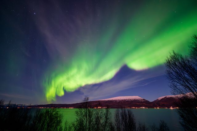 Broad aurora curtains opening above a snow-covered mountain ridge and dark shoreline.