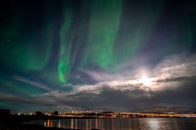 Tall green aurora curtains above a bright moon, calm water, and the illuminated Tromsø shoreline.