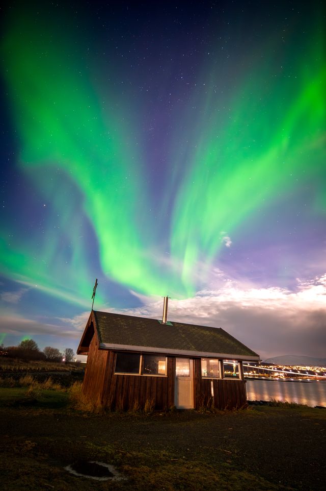 Green aurora rays fanning behind a small wooden cabin beneath moonlit clouds.