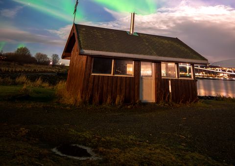 Green aurora rays fanning behind a small wooden cabin beneath moonlit clouds.