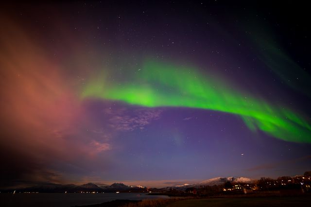 A low green aurora arc emerging through amber and violet cloud above the fjord.