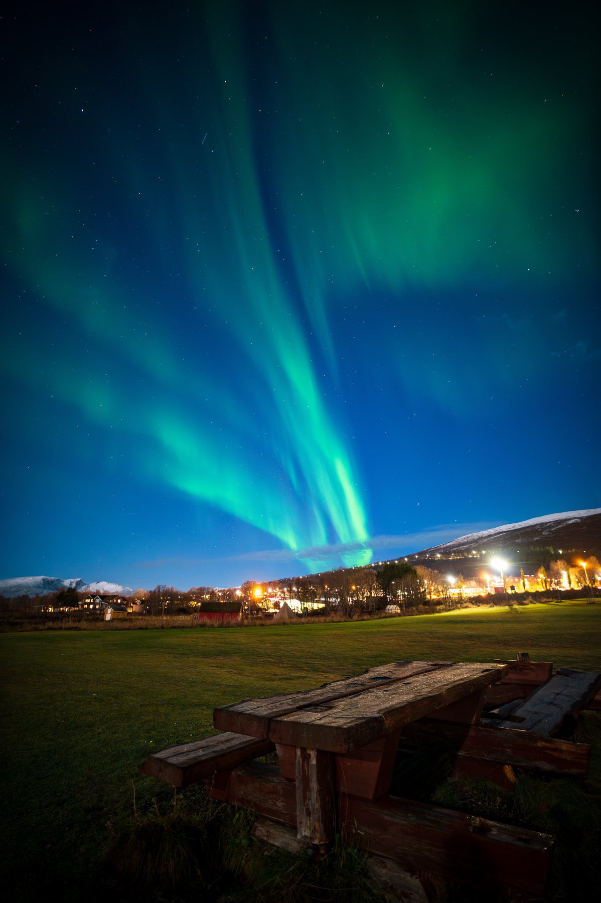 Vertical turquoise aurora beams rising behind a dark shed and a line of town lights.
