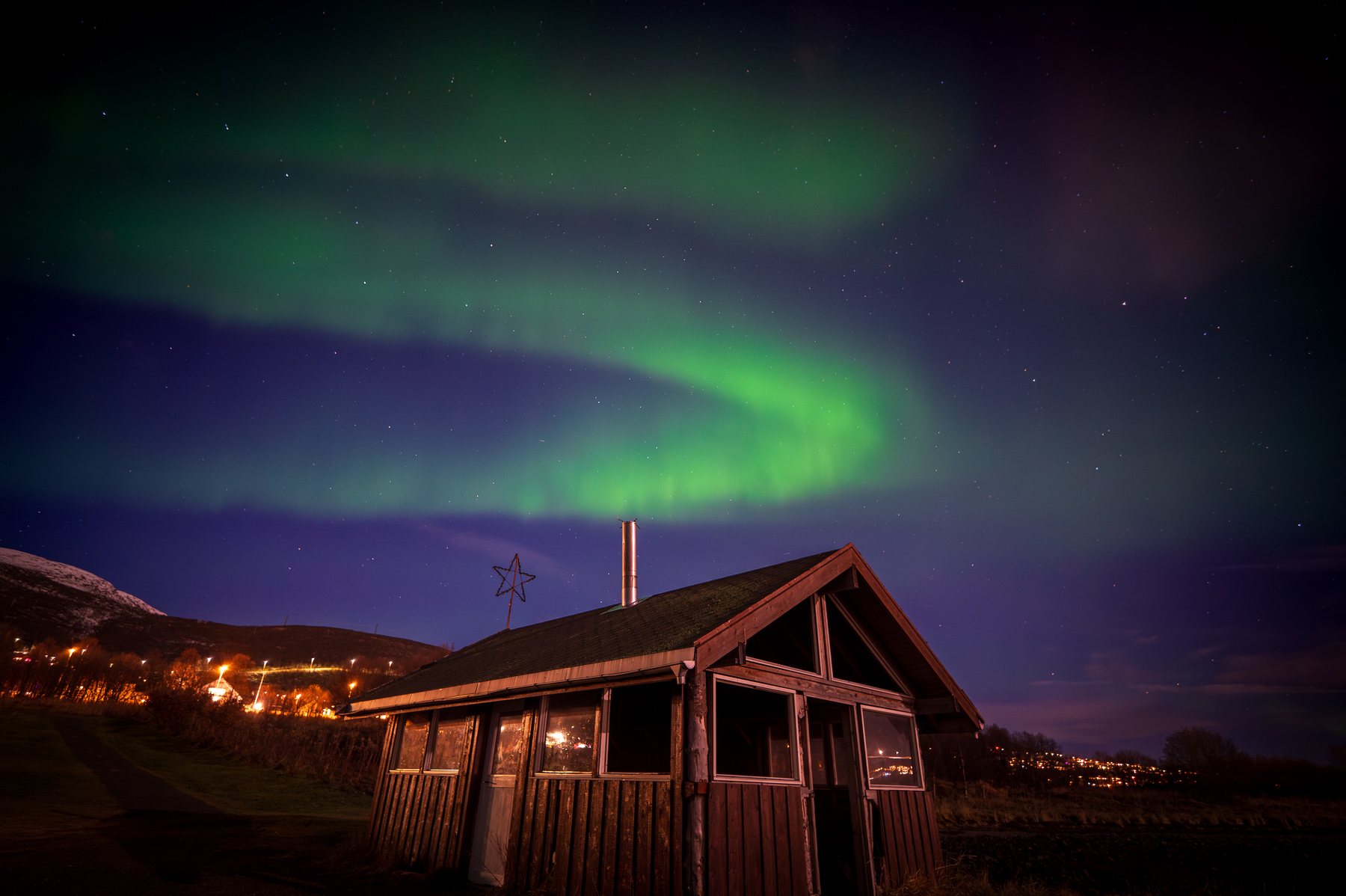 A green aurora loop suspended over a weathered wooden shed and lit Arctic hillside.