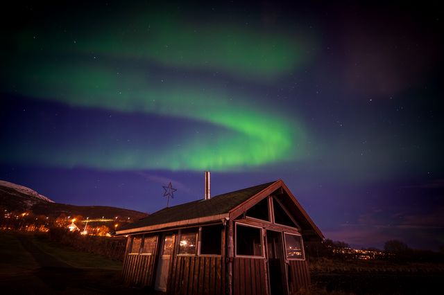 A green aurora loop suspended over a weathered wooden shed and lit Arctic hillside.