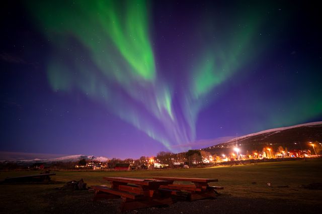 Layered green and violet aurora streaming above a village road and snow-streaked mountain.