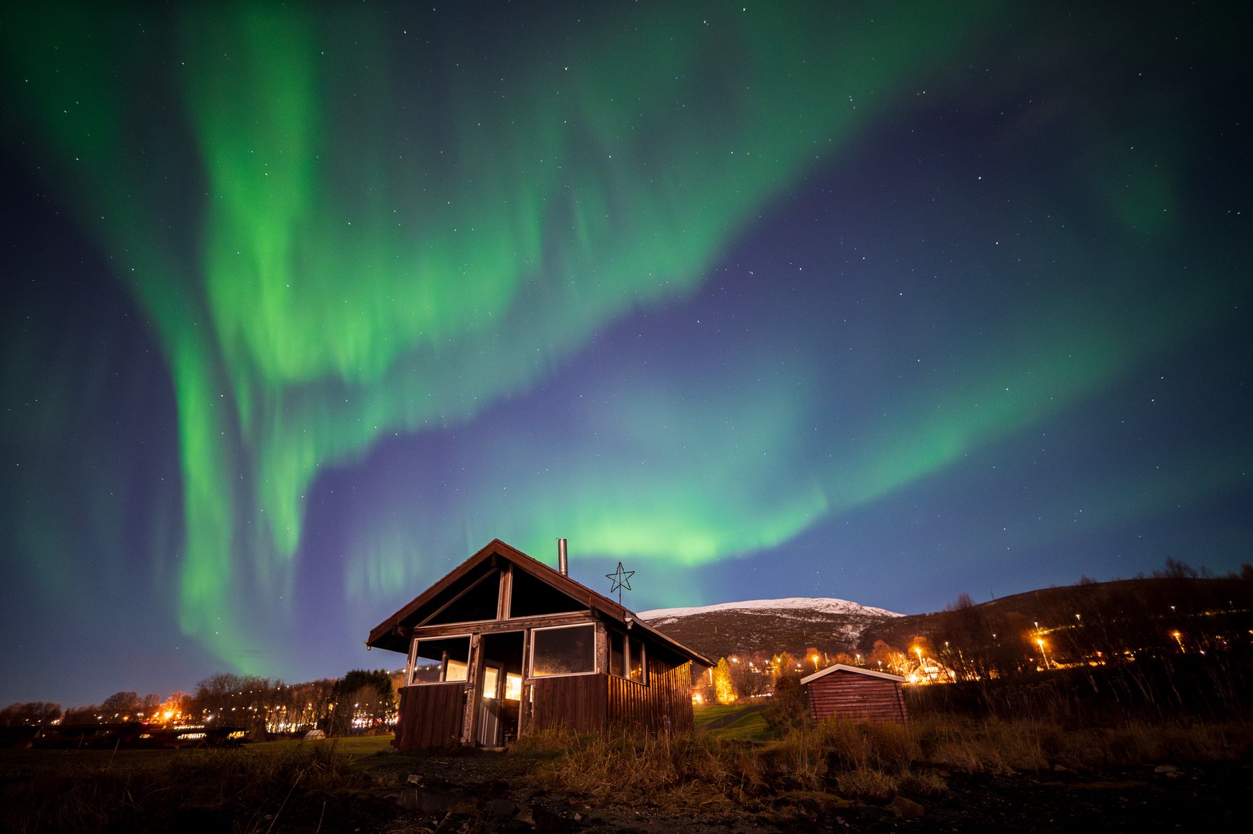 Tall green aurora curtains rising behind a dark cabin and orange shoreline lights.