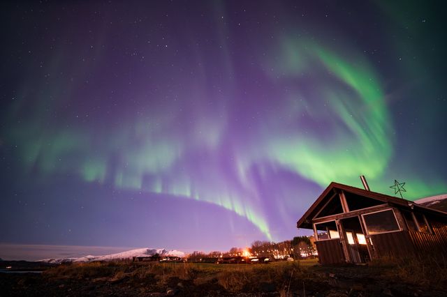 Purple and green aurora filling the sky above a wooden cabin and distant snowy ridge.