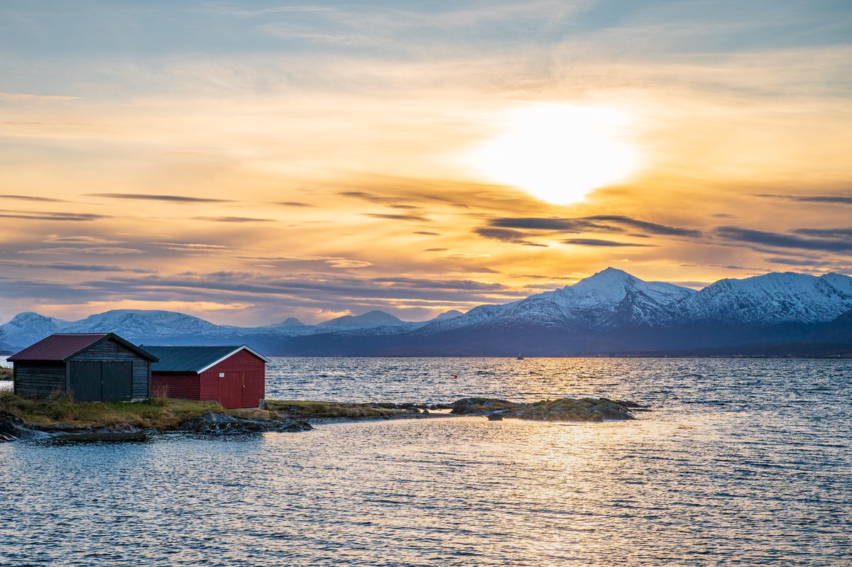 Traditional red wooden boathouses sit on a small rocky shore beneath snow-covered mountains in Northern Norway. Golden sunlight breaks through layered clouds and shimmers across the cold waters of the fjord.