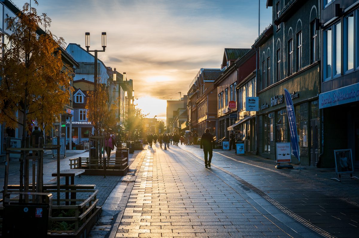 The low Arctic sun shines directly along a busy pedestrian street in central Tromsø, casting long shadows across the pavement. Autumn colours, historic wooden buildings and evening light create a warm urban scene in the heart of Northern Norway.