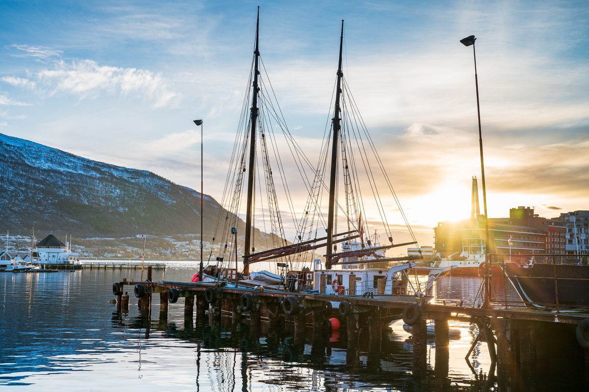 A traditional two-masted sailing vessel rests at the waterfront in Tromsø with snow-covered mountains rising behind the harbour. Bright Arctic sunlight reflects from the calm water and silhouettes the ship’s intricate rigging.