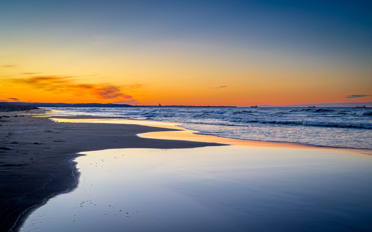 Warm orange light spreads across the horizon as evening settles over the Baltic Sea. Reflections on the wet sand mirror the fading sky while gentle waves roll toward the quiet shoreline.