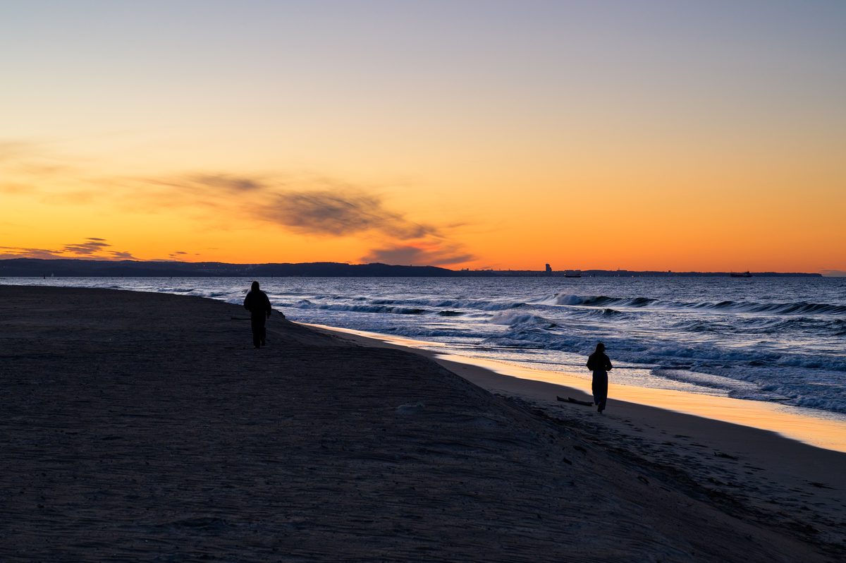 Two solitary figures walk along the Baltic shoreline as the sky burns orange above the horizon. Their silhouettes add a quiet human presence to the windswept beach and restless evening sea.
